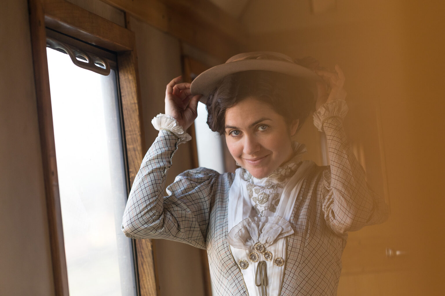 Eine Schauspielerin in viktorianischem Gewandt und mit Hut auf dem Kopf lächelt in die Kamera. Sie trägt etwas kariertes mit Rüchen, und steht vermutlich im Wohnwagen.