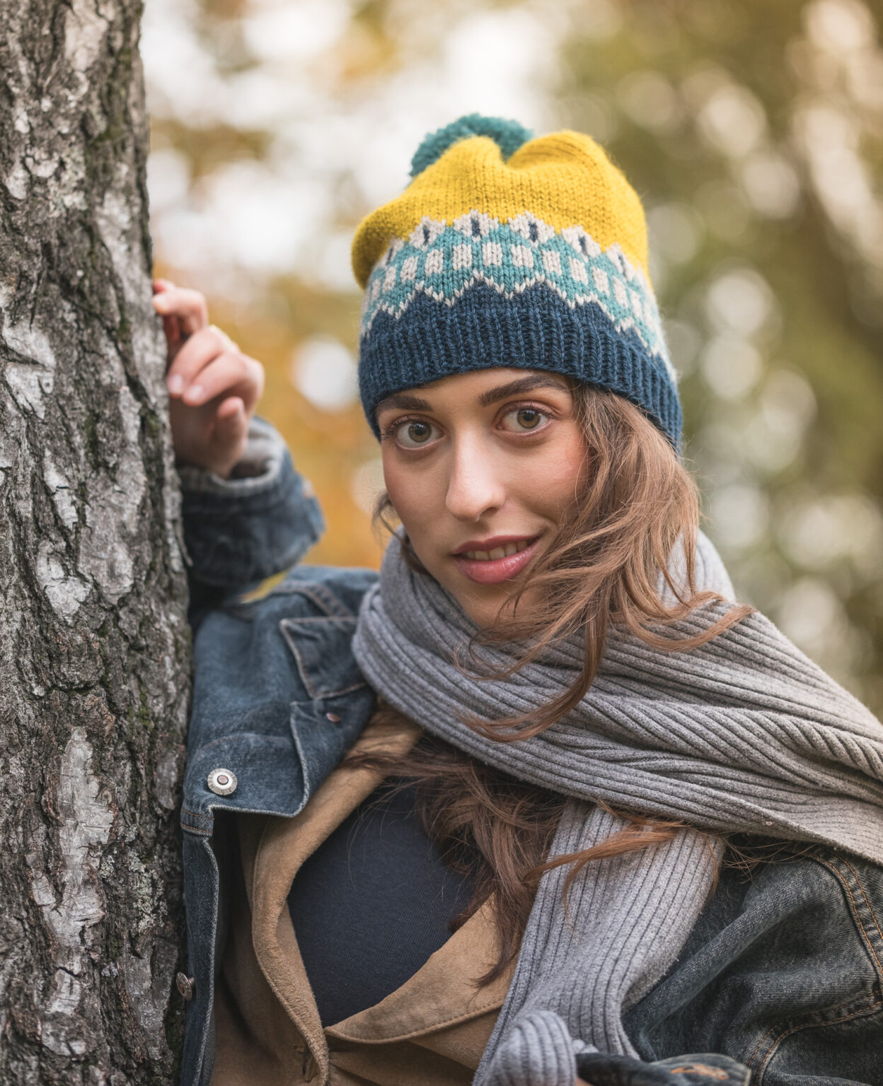 Portrait einer Frau mit braunen Haaren, großen, braunen Augen die lächelt. Sie trägt eine blau-gelb-türkise Mütze mit einem geometrischem Strickmuster, einen grauen Strickschal, eine Jeansjacke, eine braune Jacke und ein blaues Shirt darunter. Sie lehnt an einem Baum. Der Hintergrund ist unscharf und zeigt Blätter.