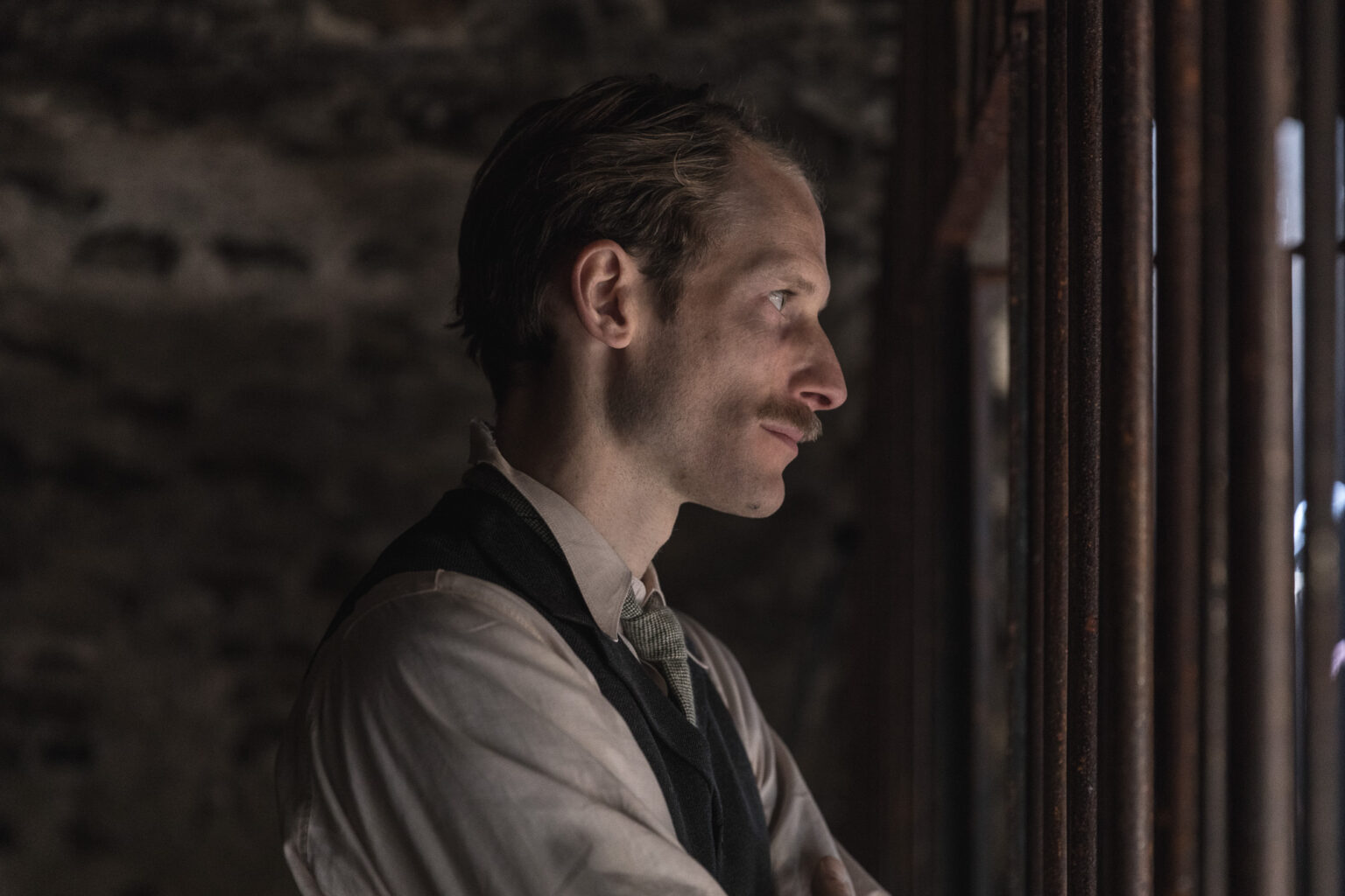 Ein Mann, der im Halbportrait seitlich im Profil fotografiert wurde, steht in einer Filmszene in einem Oldschoold Gefängnis. Er schaut durch die Gitterstäbe durch. Er hat Blonde Haare, einen Schnäuzer, ein Beiges Hemd, eine grüne Krawatte und eine schwarze Weste.
