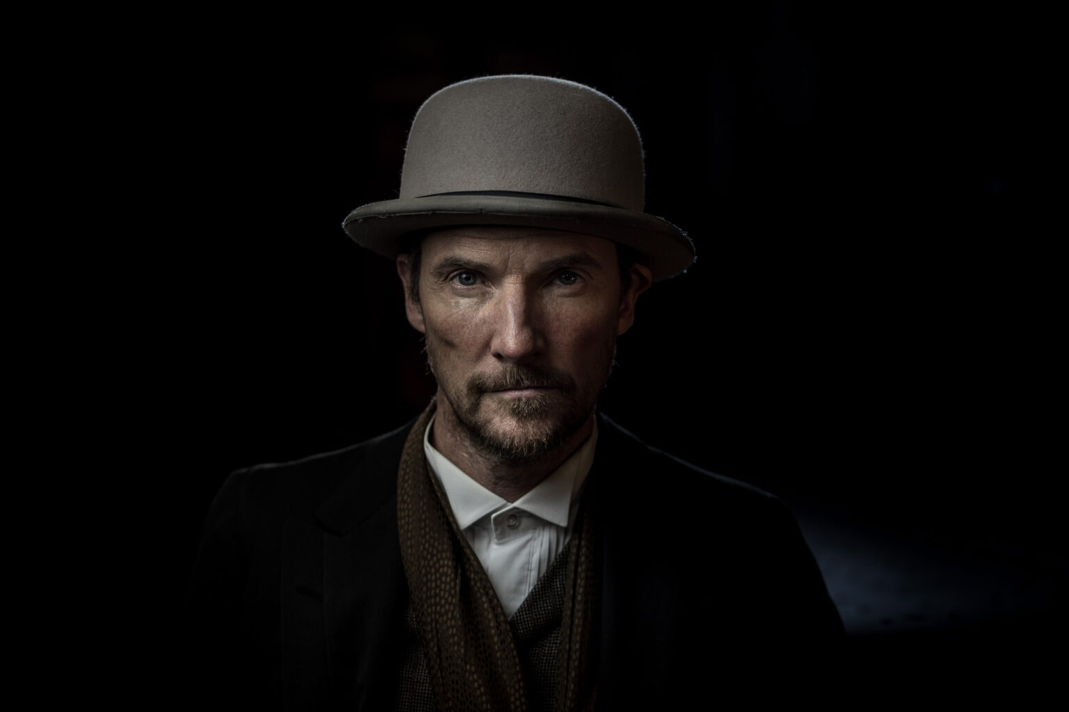 Portraitfoto mit düsterem Hintergrund, konturiertem Licht trägt eine Melone, ein Jacket, ein weißes Hemd und einen Schal. Er schaut den Betrachter direkt an.