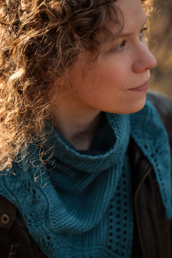 Portraitfoto einer Frau mit blonden kleinen Locken. Sie schaut nach rechts und trägt einen petrolfarbenen Strickschal mit Lochmuster und eine schwarze Lederjacke.