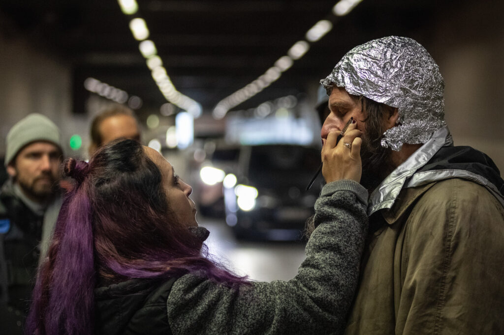Eine Frau mit lila färbenden Haaren schminkt einen Mann mit Alufolie auf dem Kopf. Im Hintergrund sieht man einen Autobahntunnel in der Unschärfe. Man sieht zudem auch zwei Männer hinter der Frau. Es scheint kühler zu sein.