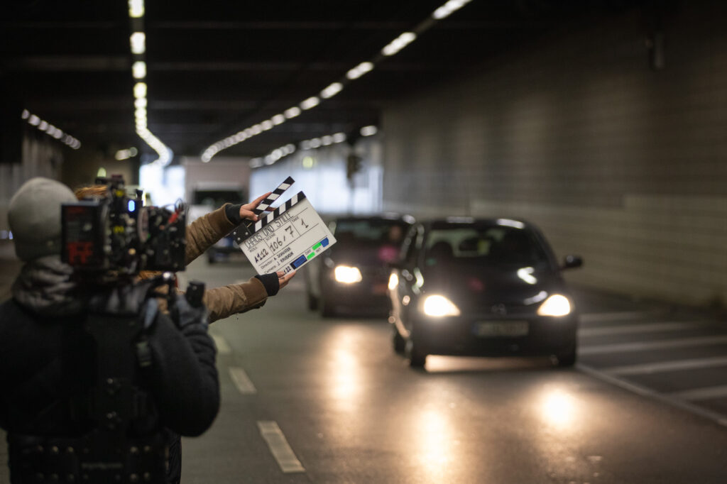 Man sieht in der Unschärfe drei Autos, die durch einen Autobahntunnel fahren. Links steht eine Person mit Kamera, dahinter eine mit einer Filmklappe. Es handelt sich hier bei um Scene 106,7 Take 1.