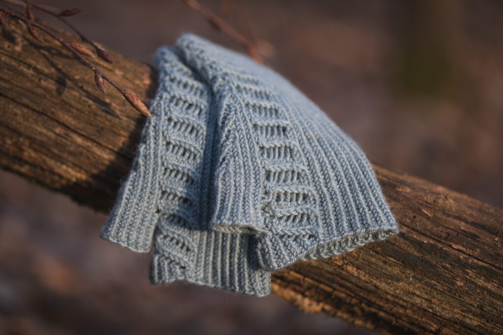 Blaue, gestrickte Stulpen liegen auf einem Baumstamm. Im Hintergrund ist Unschärfe in bräunlichen und grünen Tönen zu sehen.