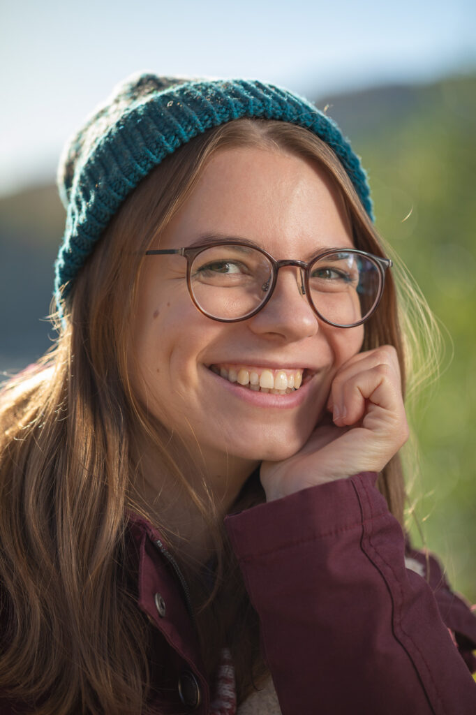Portrait einer Frau mit strahlendem Lachen, blonden Haaren, einer Brille trägt eine blaue Strickmütze und eine rote Jacke. Der Hintergrund ist grün und verschwindet in der Unschärfe.