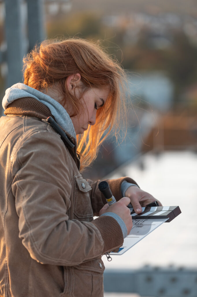 Frau mit roten Haaren und brauner Jacke, wie auch grauem Hoodie wird von hinten von der Sonne beleuchtet. Sie steht seitlich und hält einen dicken Marker und eine Filmklappe in der Hand. Sie beschriftet die Filmklappe.