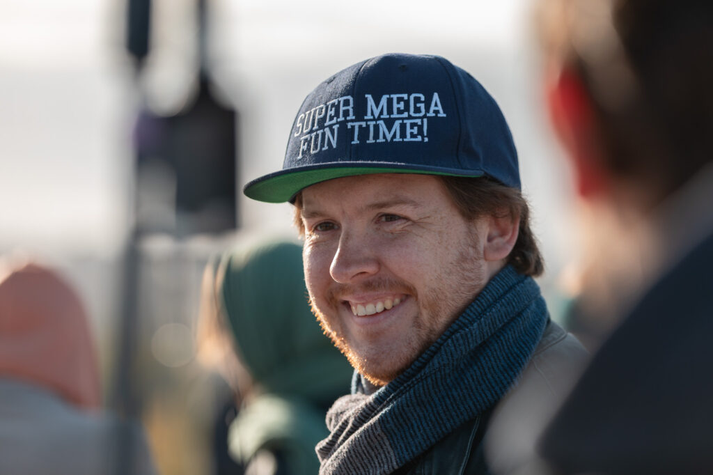 Filmcrew Mitglied lächelt jemanden an. Er ist im Halbprofil zu sehen. Er trägt eine graue Jacke einen grau-blauen Schal und eine Blau-grüne Cap mit der Aufschrift: Super mega fun time! Der Vorder-und Hintergrund sind unscharf.