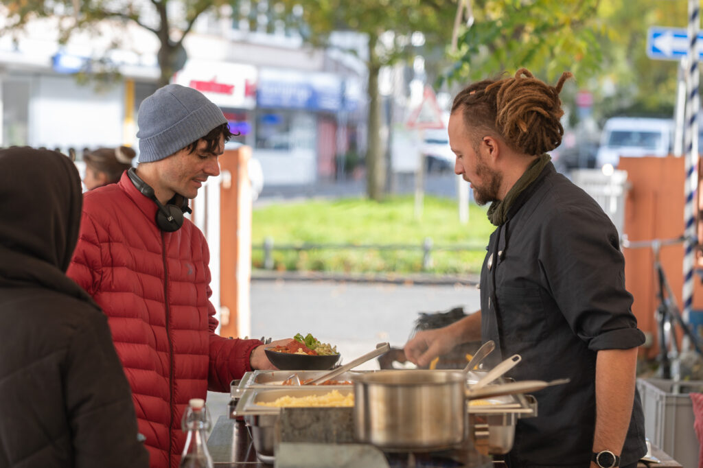 Ein Caterer mit grauer Kochkleidung steht am Buffet und bedient einen Mann mit roter Jacke, Kopfhörern, grauer Mütze. Der Hintergrund sieht sonnig aus. Die Menschen sind aber warm angezogen. Der Mann in roter Jacke hält einen Teller mit Essen fest.