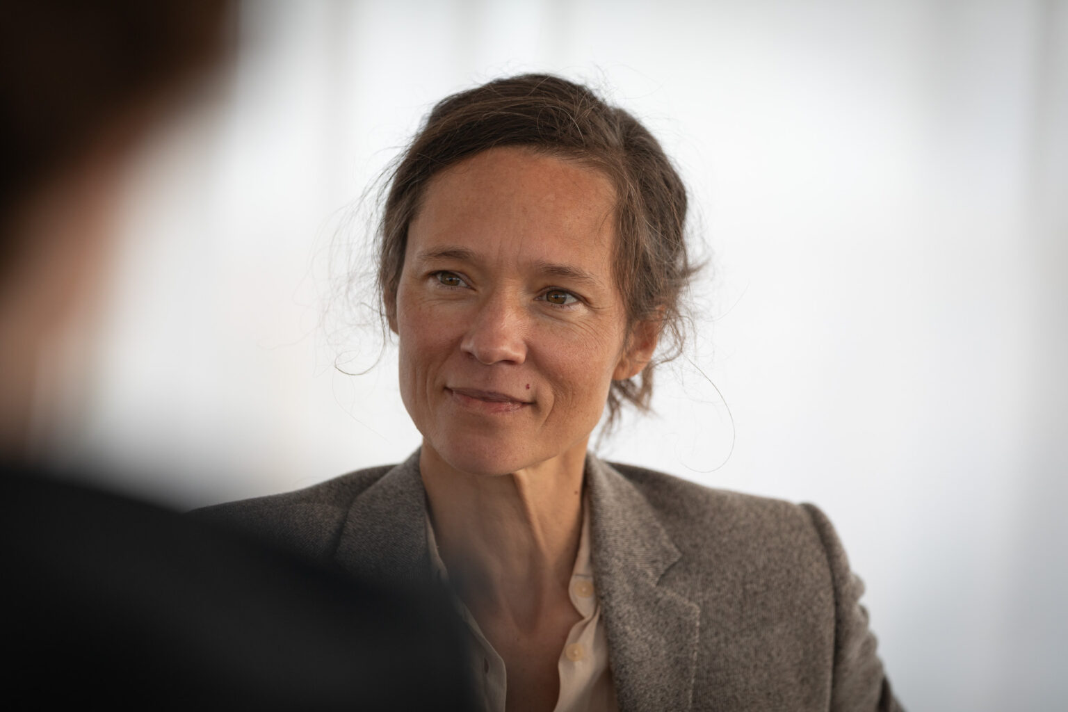 Portraitfoto eine Frau mit braunen Augen, zusammengebundenen brauenden Haaren. Sie trägt eine graue Jacke und ein rosé färbendes Hemd. Der Hintergrund ist weiß. Sie schaut auf eine Person im Vordergrund, die stark angeschnitten und unscharf ist.