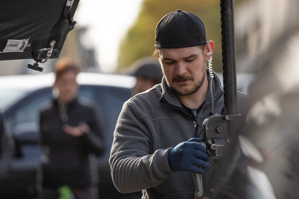 Ein Mann mit schwarzer Cap, grauem Pullover, blauen Handschuhen, Mikrofon Kabel hält ein Stativ fest. im Vorder- und Hintergrund sind diffuse Sachen und Menschen unscharf. Der Mann schaut nach unten.