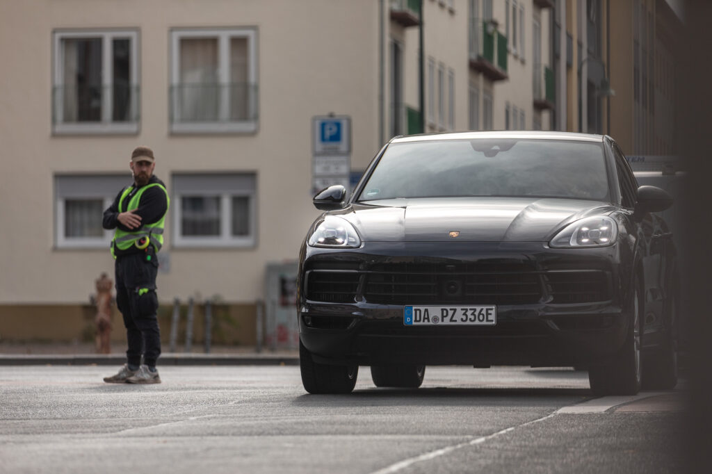 Ein Porschefahrzeug fährt auf den Betrachter zu. Es is schwarz und im Hintergrund sieht man Häuser und ein bisschen Straße. Links steht ein Mann mit brauner Cap, schwarz gekleidet mit Neongelber Sicherheitswarnweste. Er schaut auf das Auto.