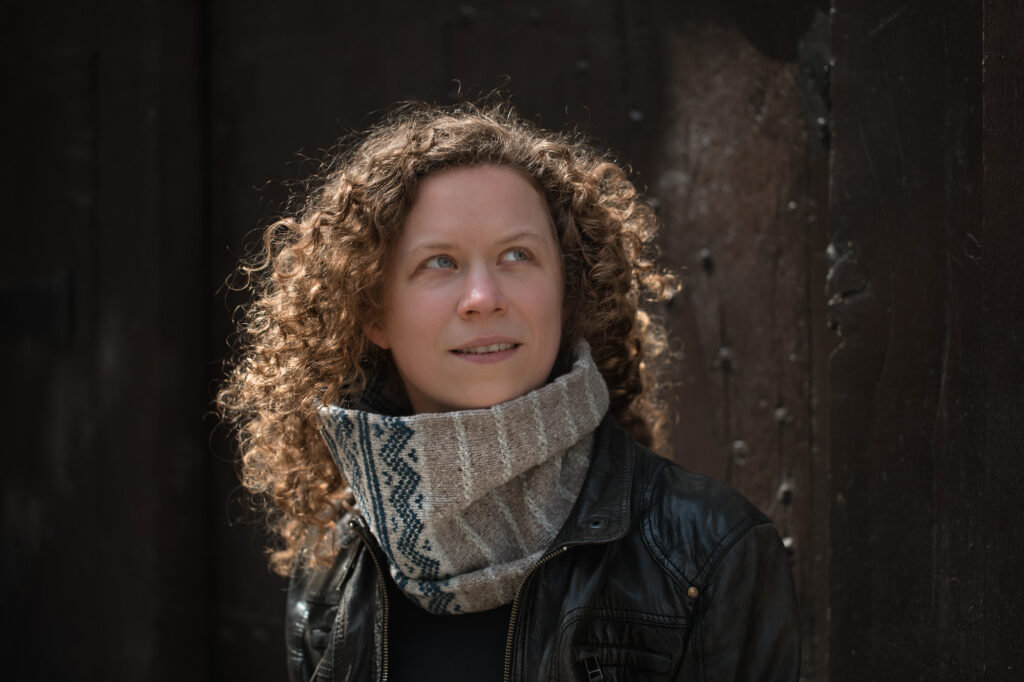 Portrait einer Frau mit blonden kleinen Locken, blauen Augen und einer Lederjacke, wie auch einem Strickschal.