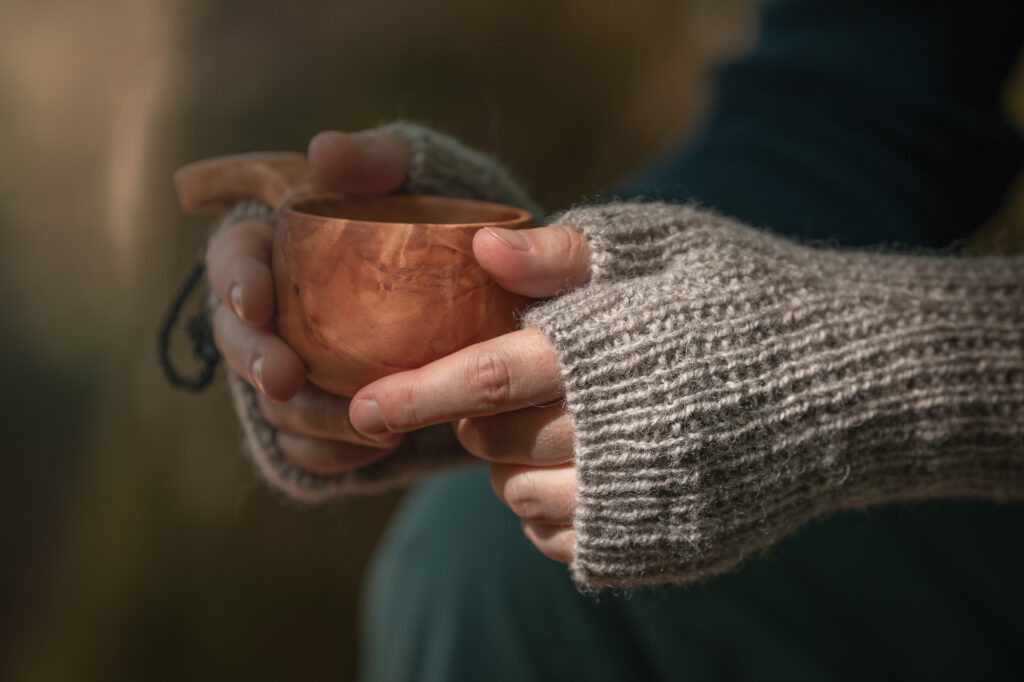 Graue, gestrickte Stulpen sind auf Händen, die ein Holzgefäß mit Henkel halten zu sehen. Die Hände sind im Close up fotografiert. Im Hintergrund sieht man in der Unschärfe ein Bein mit blauer Hose und einen Arm mit blauem Pullover.